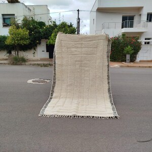May include: A beige rectangular rug with a geometric pattern and fringe detail. The rug has a textured surface and dark trim along the edges. It is displayed outdoors on a paved surface.