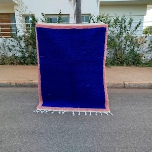 Puede incluir: Una alfombra azul y rosa tejida a mano con un borde de flecos. La alfombra está doblada por la mitad y de pie sobre una superficie pavimentada.