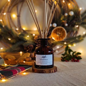 May include: A brown glass bottle with a label that reads "Gingerbread La Apothecary Reed Diffuser". The bottle is filled with a clear liquid and has wooden sticks sticking out of the top. The bottle is sitting on a wooden base. The background is blurry and features a Christmas wreath with lights.