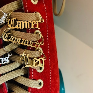 May include: Close-up of a red suede roller skate with beige laces. Gold-colored charms with the zodiac signs Cancer, Capricorn, and Leo are attached to the laces. The skate has white stitching.