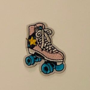 May include: An embroidered patch in the shape of a high-top roller skate. The skate is light pink with white laces, a yellow star detail on the side, and four bright blue wheels. The entire design is outlined in white thread on a fabric backing.