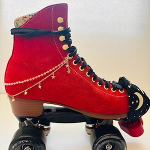 May include: A bright red suede roller skate featuring black laces, a black toe guard with a white moon and star pattern, and a gold-toned chain accessory adorned with small star charms draped along the side of the boot. The skate has black wheels.