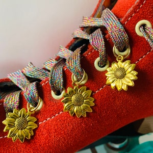 May include: Close-up of a red suede roller skate with rainbow-colored laces and gold sunflower charms. The skate has cream-colored eyelets and white stitching. The sunflower charms are hanging from the laces.