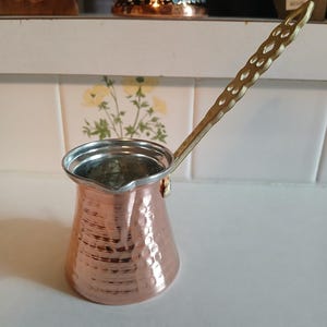 May include: A copper-colored Turkish coffee pot with a long, decorative handle. The pot has a hammered texture and a silver-colored rim. The handle is gold-toned with a patterned design. The pot is designed for brewing coffee.