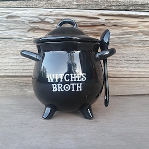 May include: A black ceramic cauldron with a lid, handles, and three feet. The words "WITCHES BROTH" are printed in white on the front. A matching black spoon rests beside the cauldron. The cauldron is a decorative item.