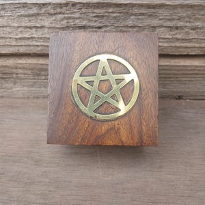 May include: A square wooden box with a brass pentagram emblem. The box is made of dark brown wood with visible grain. The pentagram is centered on the top surface and is a bright gold color.