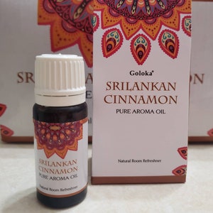 May include: A small brown glass bottle with a white cap, labeled "Sri Lankan Cinnamon Pure Aroma Oil" and "Natural Room Refreshner". The bottle is in front of a box with the same label, featuring a colorful mandala design.