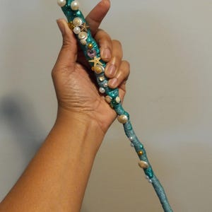 May include: A teal-colored wand with a gold seashell top, adorned with pearls, seashells, and starfishes. The wand is wrapped in teal thread and decorated with various sea-themed embellishments. The wand is held in a hand.