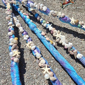 Peut inclure: Quatre baguettes décoratives enveloppées dans un matériau texturé bleu et violet, ornées de divers coquillages, de petites étoiles de mer blanches et de perles blanches rondes. Les baguettes présentent un thème côtier cohérent.