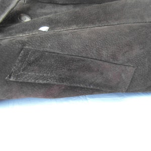 May include: Close-up of a black suede jacket with a flap pocket on the left side. The pocket has a stitched edge and is slightly angled.