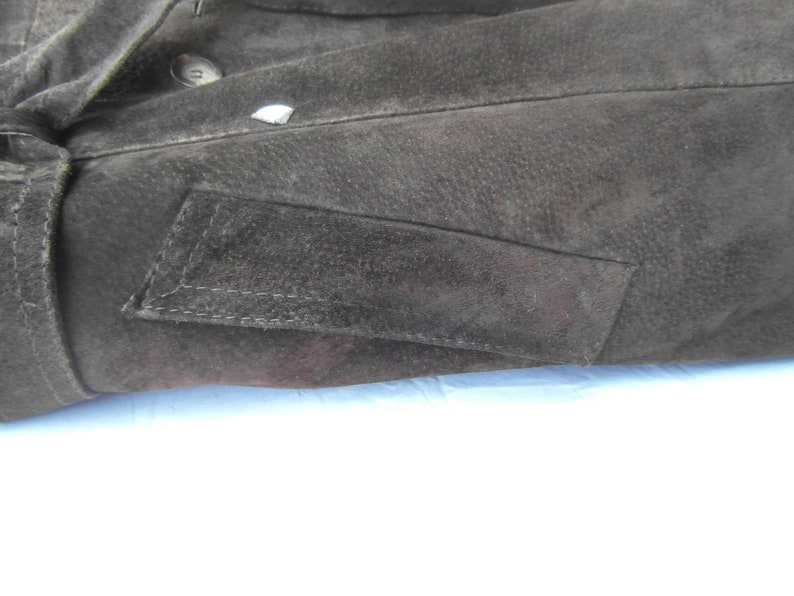 May include: Close-up of a black suede jacket with a flap pocket on the left side. The pocket has a stitched edge and is slightly angled.