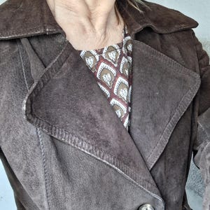May include: A brown suede jacket with a double-breasted closure and a belt. The jacket has a collar and two pockets.