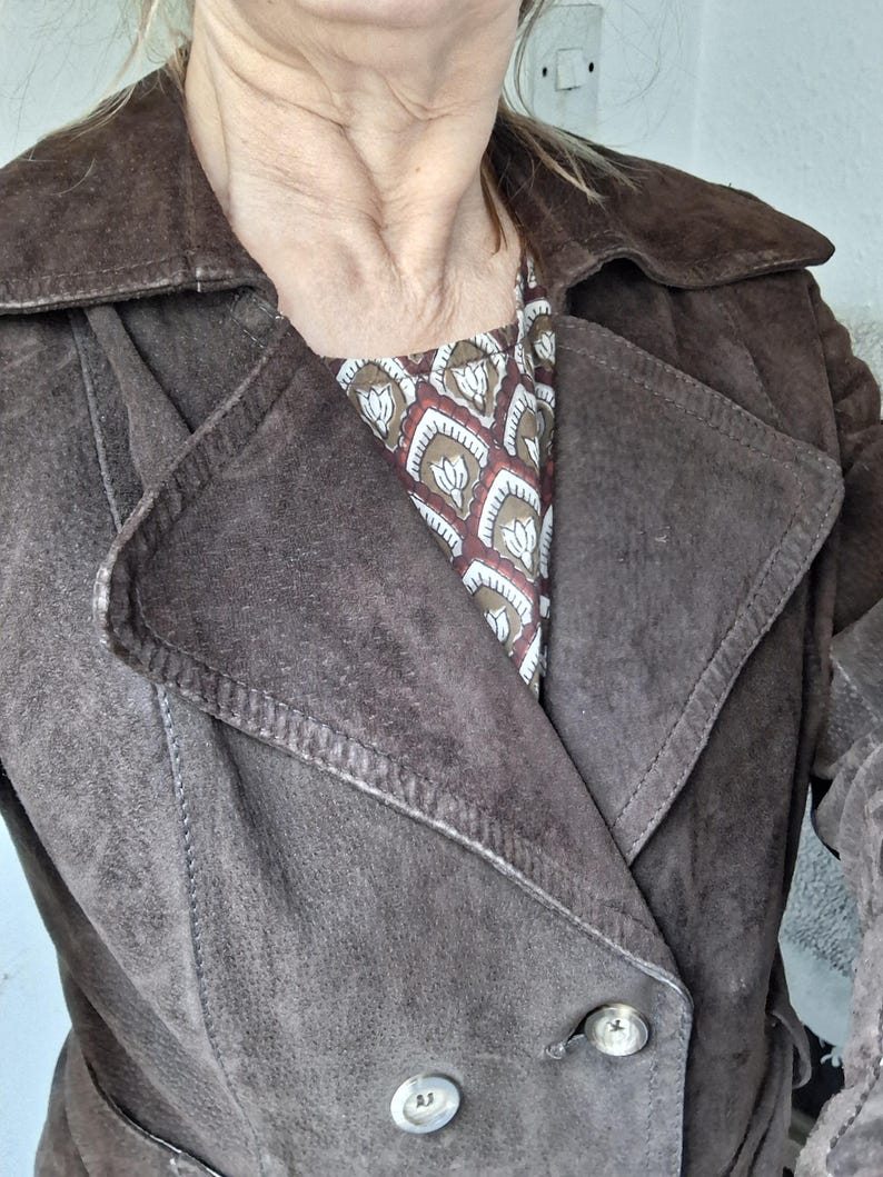 May include: A brown suede jacket with a double-breasted closure and a belt. The jacket has a collar and two pockets.