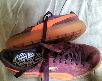 May include: Burgundy Puma Suede platform trainers with orange detailing. The trainers feature thick, light brown soles and burgundy laces. The Puma logo is visible on the side and insole.