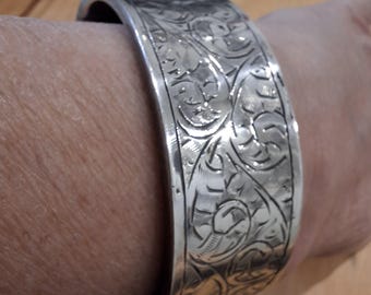 May include: A wide, silver-colored cuff bracelet with an engraved floral pattern. The bracelet is hinged and appears to be made of metal. The intricate design covers the entire surface of the bracelet, creating a vintage or antique aesthetic.