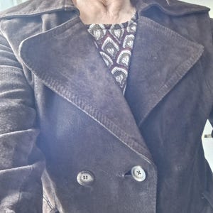 May include: A brown suede trench coat with a double breasted closure and a belt. The coat has two large buttons and a collar.