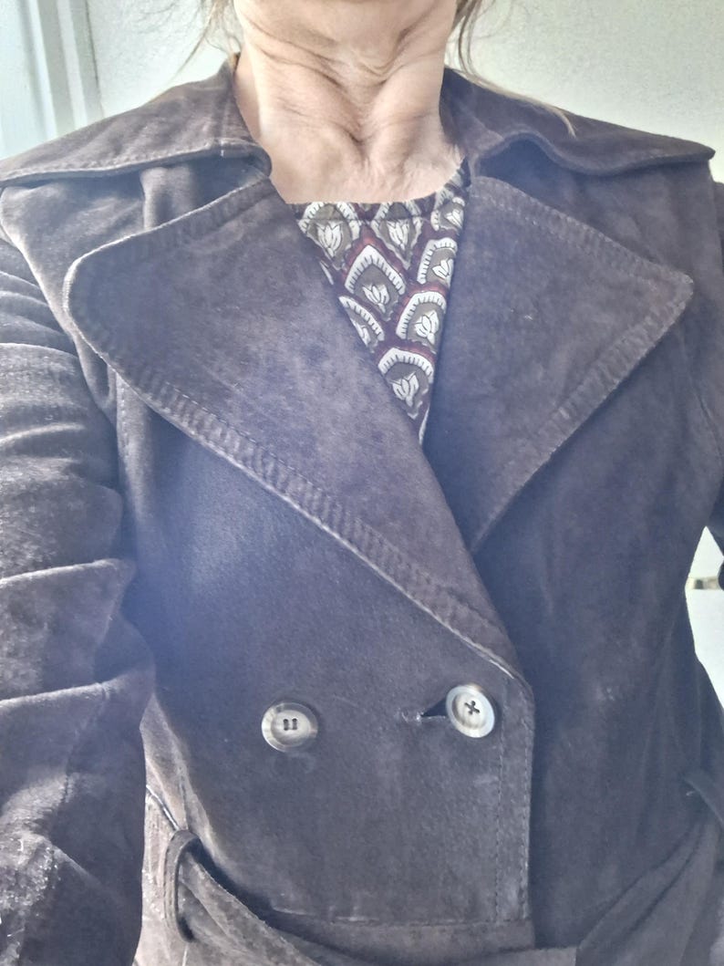 May include: A brown suede trench coat with a double breasted closure and a belt. The coat has two large buttons and a collar.