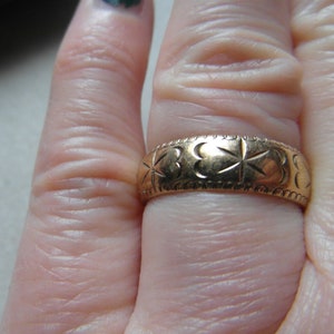 May include: A gold band ring with a clover design. The ring is worn on a finger.