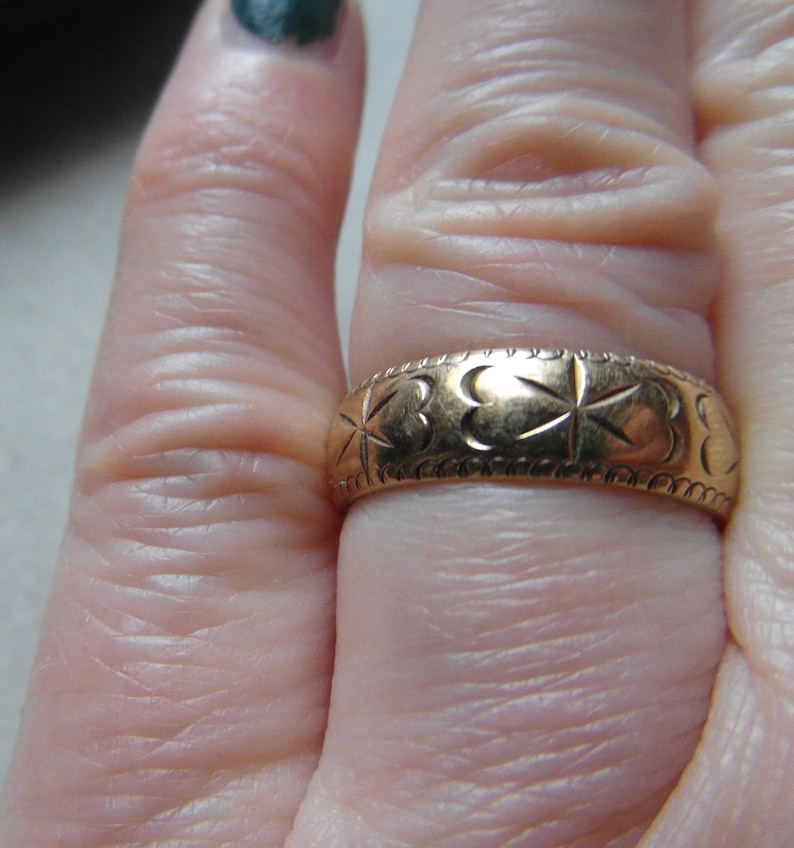 May include: A gold band ring with a clover design. The ring is worn on a finger.