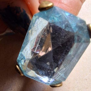 Può includere: Una gemma sfaccettata, azzurra, di forma geometrica, forse un ciondolo o un elemento decorativo. La gemma è tenuta in posizione da quattro piccole clip metalliche color oro. La pietra presenta crepe interne.