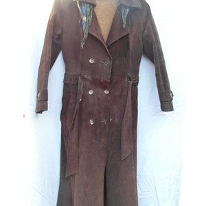 May include: A long brown suede coat with a double breasted closure and a belt. The coat has a collar and is hanging on a hanger.