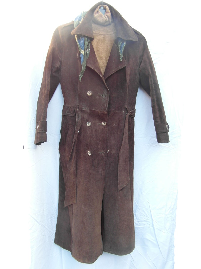 May include: A long brown suede coat with a double breasted closure and a belt. The coat has a collar and is hanging on a hanger.