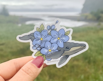 Può includere: Un adesivo raffigurante una balena grigia con fiori azzurri e foglie verdi. La balena ha un ventre bianco e un piccolo occhio. L'adesivo è tenuto davanti a uno sfondo sfocato all'aperto.