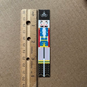 May include: A colorful nutcracker figurine with a black hat, blue jacket, white shirt, red pants, and black boots. The figurine is approximately 3 inches tall.