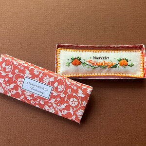 以下が含まれることがあります： オレンジと白の花柄の長方形の箱と、刺繍された作品。 刺繍には、オレンジ色のカボチャと緑のつるで「HARVEST Blessings」という言葉が描かれています。 箱には「Chelsea Little & Co. Embroidery」と書かれています。