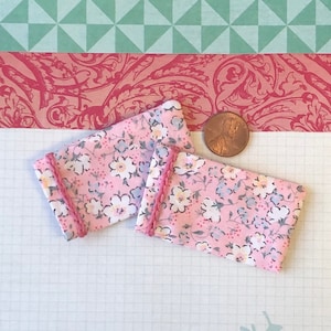 Può includere: Due piccoli cuscini in tessuto rosa con stampa floreale e bordino rosa. I cuscini sono su uno sfondo bianco a griglia.