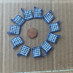 Puede incluir: Una colección de mini agarraderas cuadradas, dispuestas en círculo, cada una con un patrón de cuadros azules y blancos y un lazo azul. Una moneda de un centavo está en el centro. Las agarraderas son de tela.