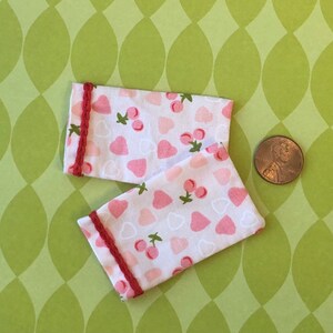 Può includere: Due polsini in tessuto bianco con bordino rosso e un motivo a cuori e ciliegie rosa e bianche.