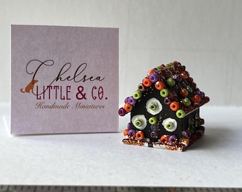 May include: A miniature Halloween gingerbread house decorated with black, orange, green, and purple beads. The house has a black base with white accents and a glitter finish. The roof is decorated with a variety of beads and has a gingerbread look. The house is about 2 inches tall.