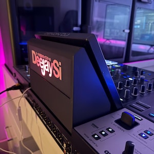 May include: A black Denon DJ controller featuring a prominent screen with the illuminated text DeejaySi in orange and white. The console includes a silver control panel with Serato branding, various knobs, buttons, and input ports along the side.