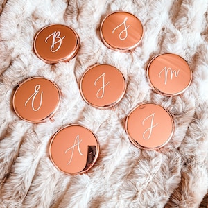 May include: Seven rose gold compact mirrors, each with a different white cursive letter: B, S, E, J, M, J, and A. They are arranged on a fluffy, light brown surface.