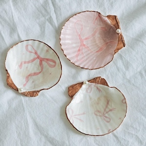 May include: Three decorative seashell dishes. Each shell is painted with a pink ribbon design and has a gold-coloured rim. One shell has a small pearl attached. The shells are on a white background.