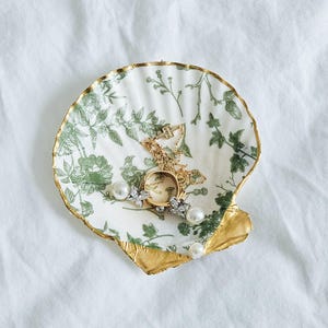 May include: A decorative seashell-shaped trinket dish with a gold-painted edge and a green floral pattern. The dish holds a gold necklace, a watch, and several pearl earrings. The background is a white fabric.