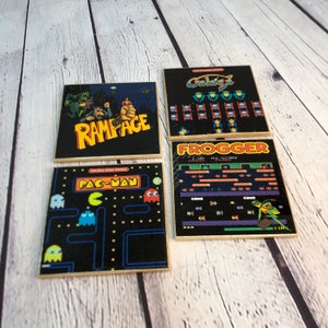 Peut inclure: Quatre sous-verres carrés avec des graphismes de jeux vidéo rétro. Les sous-verres représentent Pac-Man, Frogger, Rampage et Galaxian. Les sous-verres sont noirs avec des graphismes colorés et une bordure dorée.