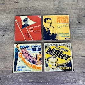 May include: Four vintage, colorful, square, metal signs featuring Glenn Miller and his orchestra. The signs feature images of Glenn Miller, a trombone, and a band. The signs have text that reads "Major Glenn Miller A.E.F. orchestra", "String of Pearls Glenn Miller", "George Montgomery Ann Rutherford Glenn Miller and his band Orchestra Wives Bari Landis Romero A 20th Century Fox Picture", and "Now Appearing in Person at Baltimore's Hippodrome Ballroom Glenn Miller and his Orchestra".