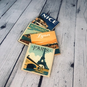 May include: Four vintage-style postcards featuring images of Paris, Lyon, Normandy, and Nice, France. Each postcard has a blue, yellow, and white color scheme with a vintage font.