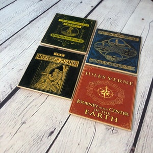 May include: Four square coasters with book cover designs. Titles include "Twenty Thousand Leagues Under the Seas", "Around the World in Eighty Days", "The Mysterious Island", and "Journey to the Center of the Earth". The coasters have a vintage aesthetic.