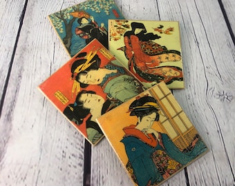 以下が含まれることがあります： 伝統的な衣装を着た女性を描いた日本の木版画が施された正方形のコースター4枚。コースターは、薄い木目調のクリーム色の背景に印刷されています。