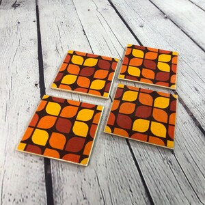May include: Set of four square coasters featuring a geometric pattern in shades of orange, brown, and yellow. The coasters have a retro 1970s style.