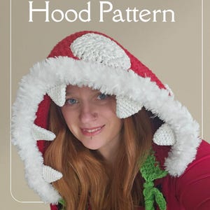 Puede incluir: Un patrón de capucha de ganchillo hecho a mano con un diseño de seta roja y blanca. La capucha presenta un ribete de piel sintética blanca y detalles verdes en forma de vid. El texto "Happy Kit Crochet" y "Man Eater Hood Pattern" son visibles.