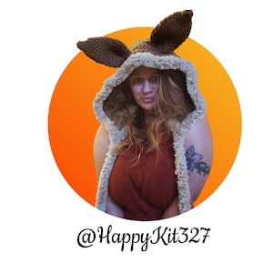 May include: A brown and beige crocheted hood with large, upright ears. The hood has a furry trim and is worn over a rust-colored top. The image is set against an orange circle with the text "@HappyKit327" at the bottom.