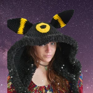 May include: A black crocheted hood featuring two tall, pointed ears with yellow horizontal stripes and a yellow circular ring emblem centered on the forehead. The hood is crafted from a soft, textured yarn and covers the head with a wide opening.