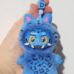 May include: A blue, plush creature figurine with a detailed face, large yellow eyes, and white fangs. The figurine has a blue hood with pink accents and a small blue loop with a letter "D" on top. The body has a leopard print.