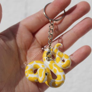 May include: A handcrafted keyring with a yellow and white snake design. The snake is made from a smooth material and features a detailed pattern. The keyring includes a silver-coloured chain and ring for attaching to keys or bags.