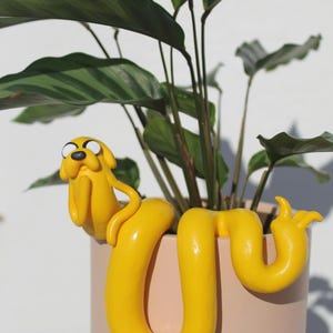 Pode incluir: Uma escultura de argila amarela de um cão de desenho animado, que se assemelha ao Jake do Hora de Aventura, está enrolada em torno de um vaso rosa com uma planta verde. O cão está sentado na borda do vaso com a cabeça inclinada para o lado.