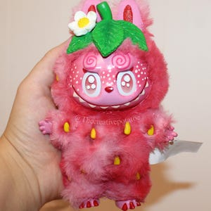 Könnte beinhalten: Ein rosa Plüschtier in Erdbeerform mit einem lächelnden Gesicht, weißen Punkten und gelben Samen. Es hat ein grünes Blatt und eine weiße Blume. Der Text "@Thecreativepotatoe" ist sichtbar.